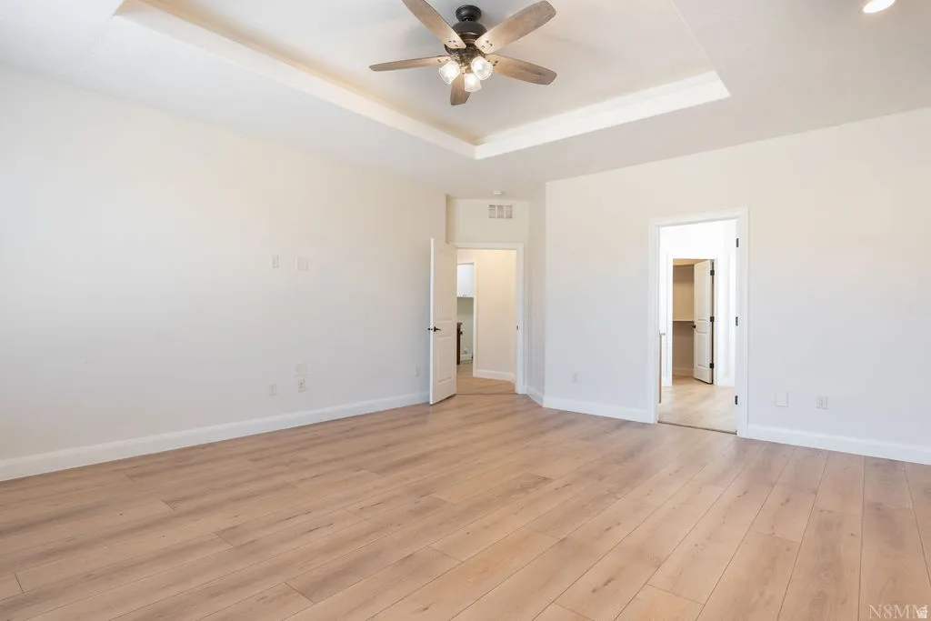 Spacious room featuring light wood-finish flooring, a tray ceiling, and a ceiling fan with integrated lighting