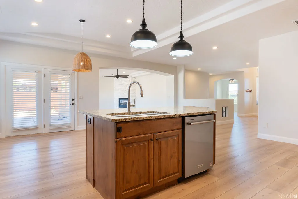 Kitchen island featuring a stainless steel sink with a high-arc faucet, granite countertop, wood cabinetry, and a built-in dishwasher