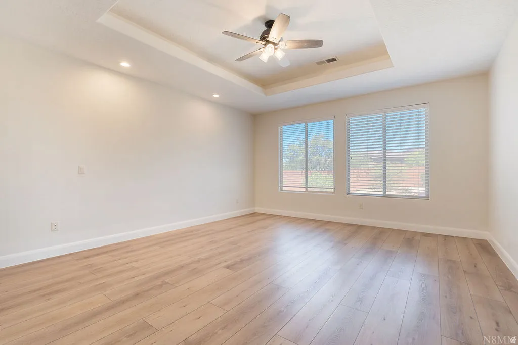 Spacious room featuring light wood-finish flooring, a tray ceiling with a ceiling fan, recessed lighting, and two windows with blinds