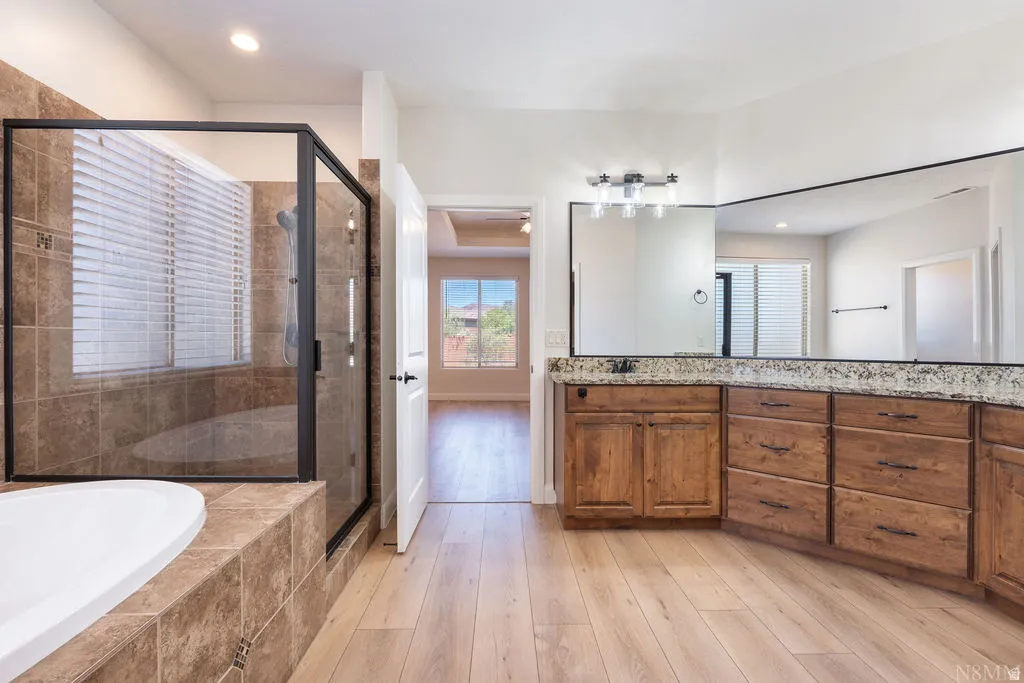 Spacious bathroom featuring a large soaking tub, separate glass-enclosed shower with tile surround, extensive wood-finish vanity with stone countertop, and light wood-finish flooring