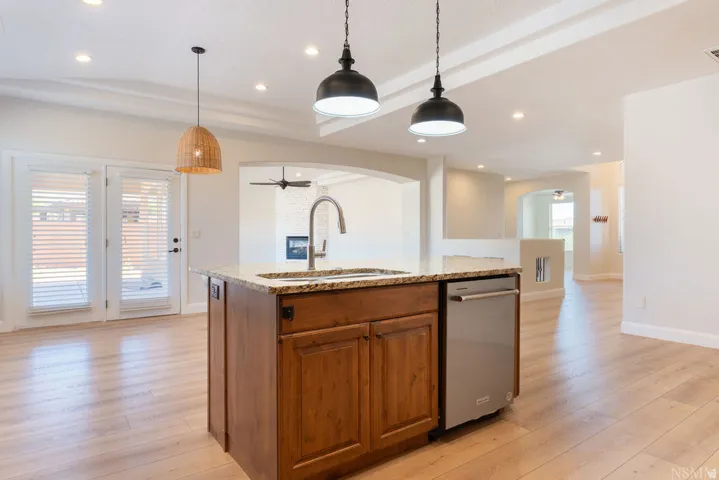 Kitchen island featuring a stainless steel sink with a high-arc faucet, granite countertop, wood cabinetry, and a built-in dishwasher