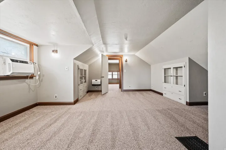 Carpeted room with sloped ceilings, featuring a built-in window unit, wall sconces, and built-in cabinetry with glass-front uppers and drawer storage