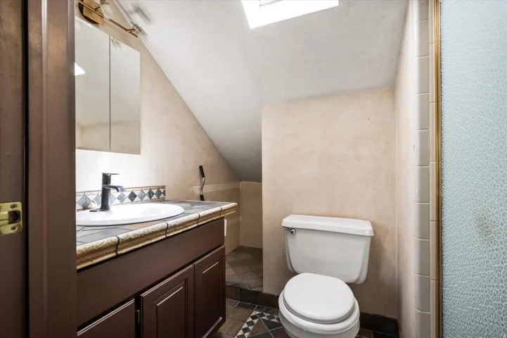 Bathroom featuring a skylight, dark wood vanity with a white basin sink, tiled countertop, and a decorative tile backsplash
