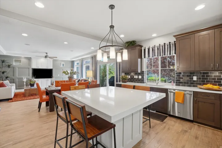 Open-concept kitchen and living area featuring wood-finish flooring, a large kitchen island with seating, dark wood cabinetry, a dark tile backsplash, and recessed lighting
