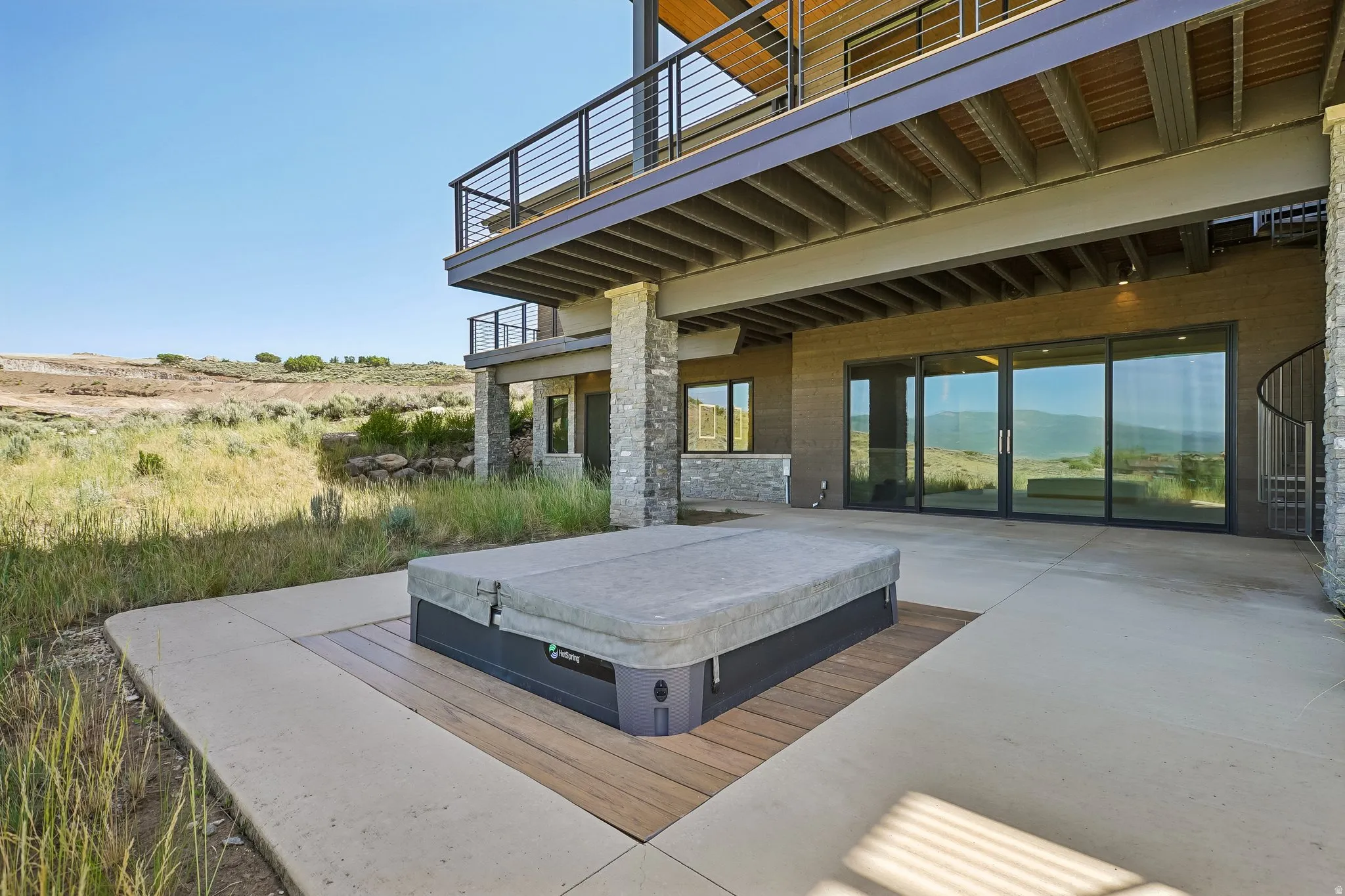 Outdoor patio featuring a built-in hot tub with a wood-finish surround, extensive concrete paving, and a stacked stone exterior wall