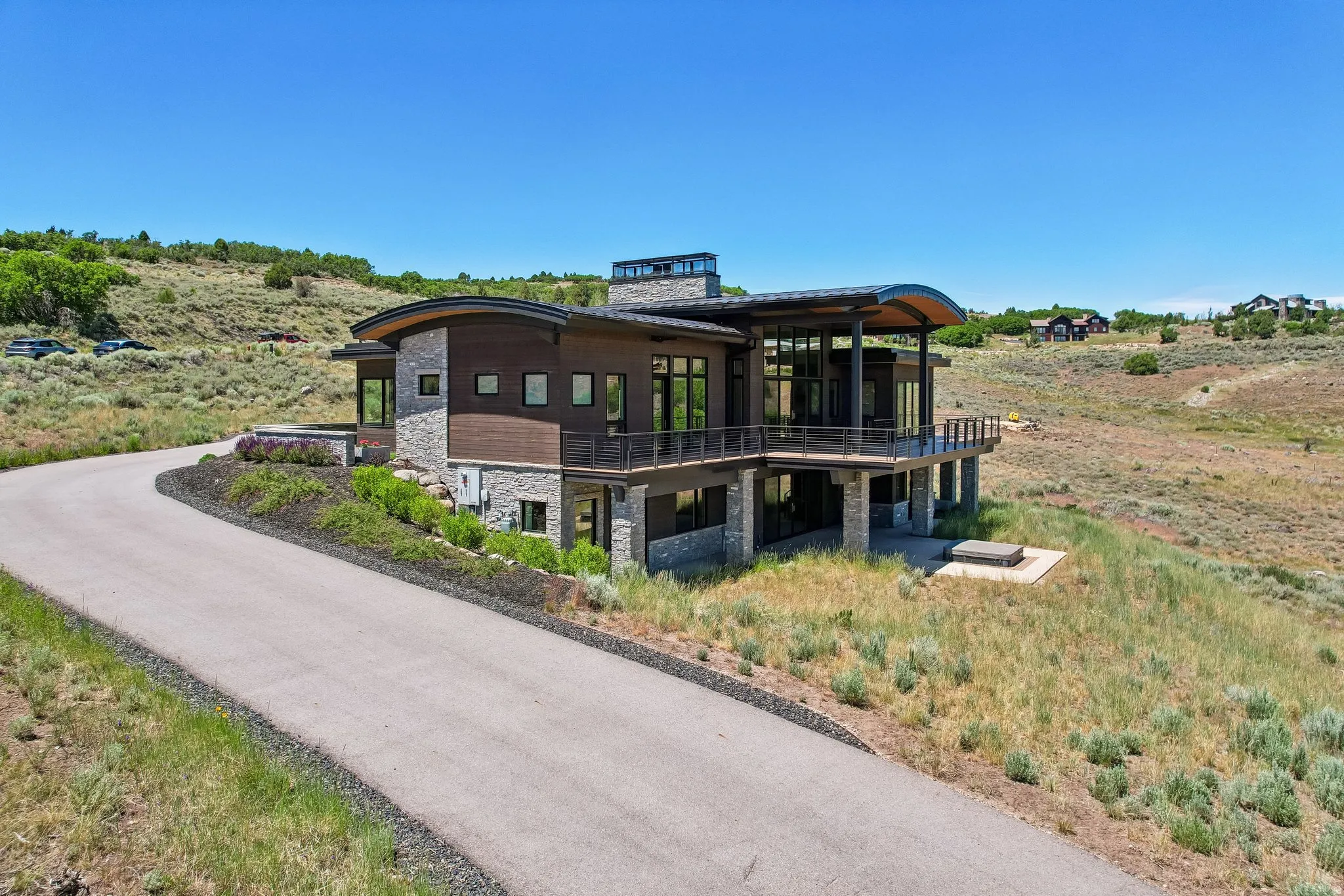 Modern architectural residence featuring stone and wood siding, a curved roofline, expansive floor-to-ceiling windows, an elevated balcony with metal railings, and a lower-level patio