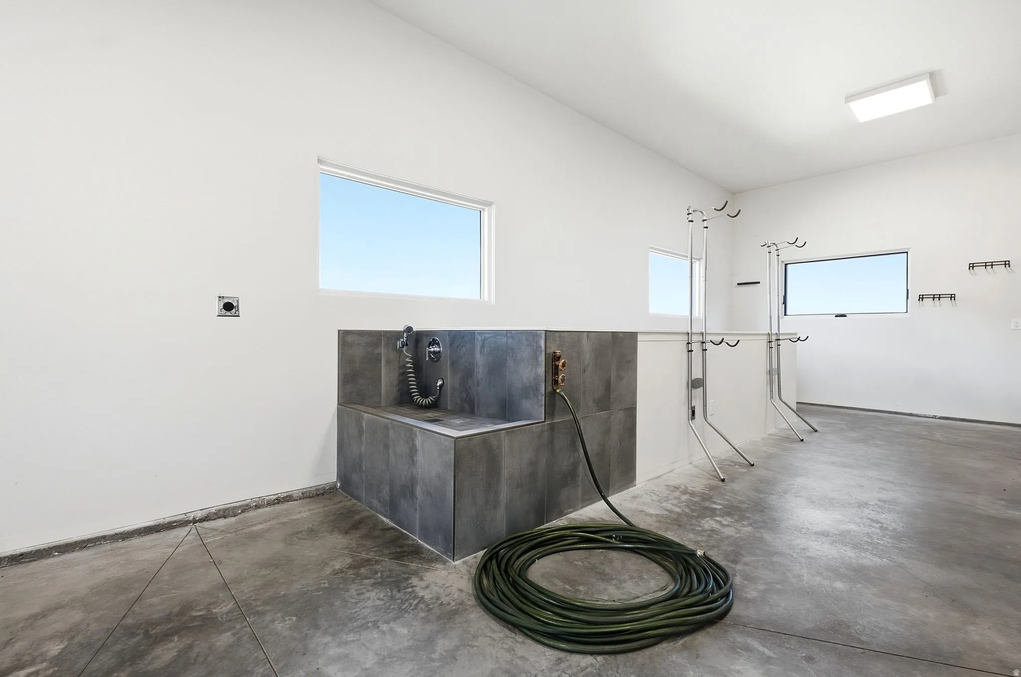 Utility sink with dark tiled surround and flexible hose faucet, concrete flooring, rectangular windows, and overhead flush-mount lighting