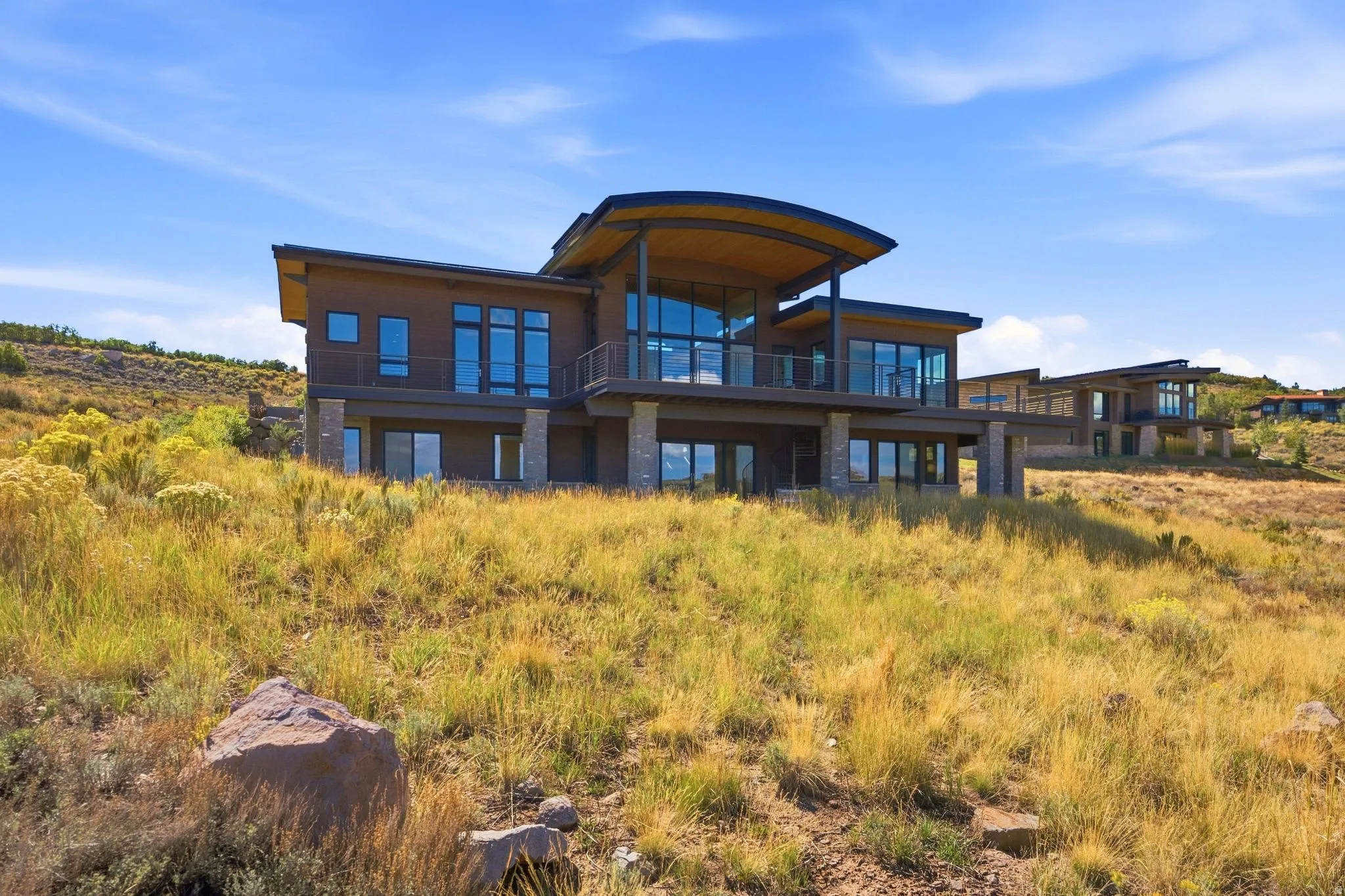 Modern architectural style with expansive windows, a curved roofline, multiple balconies, and stone-clad support columns