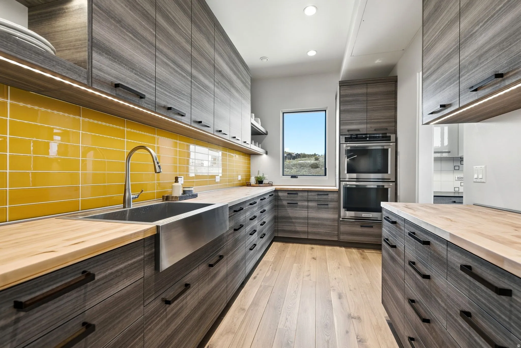 Contemporary kitchen featuring wood-finish flooring, full-height gray wood-finish cabinetry, light wood-finish countertops, a yellow subway tile backsplash, and built-in stainless steel double ovens