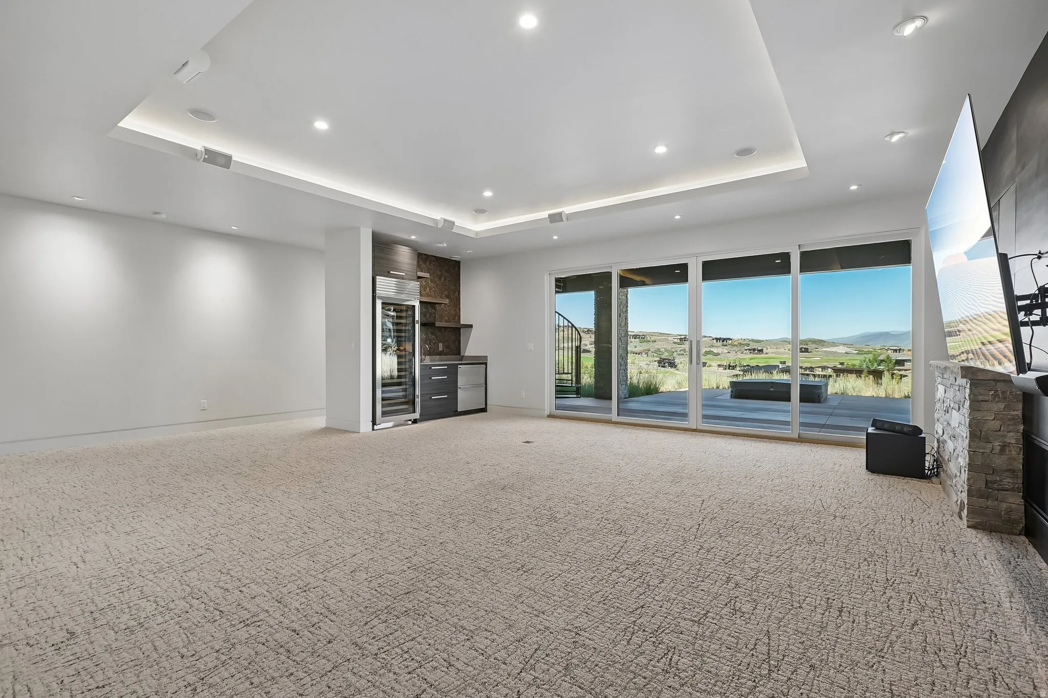 Expansive room featuring multi-panel sliding glass doors, a built-in wet bar with a wine refrigerator, a stone-clad fireplace, and recessed ceiling lighting
