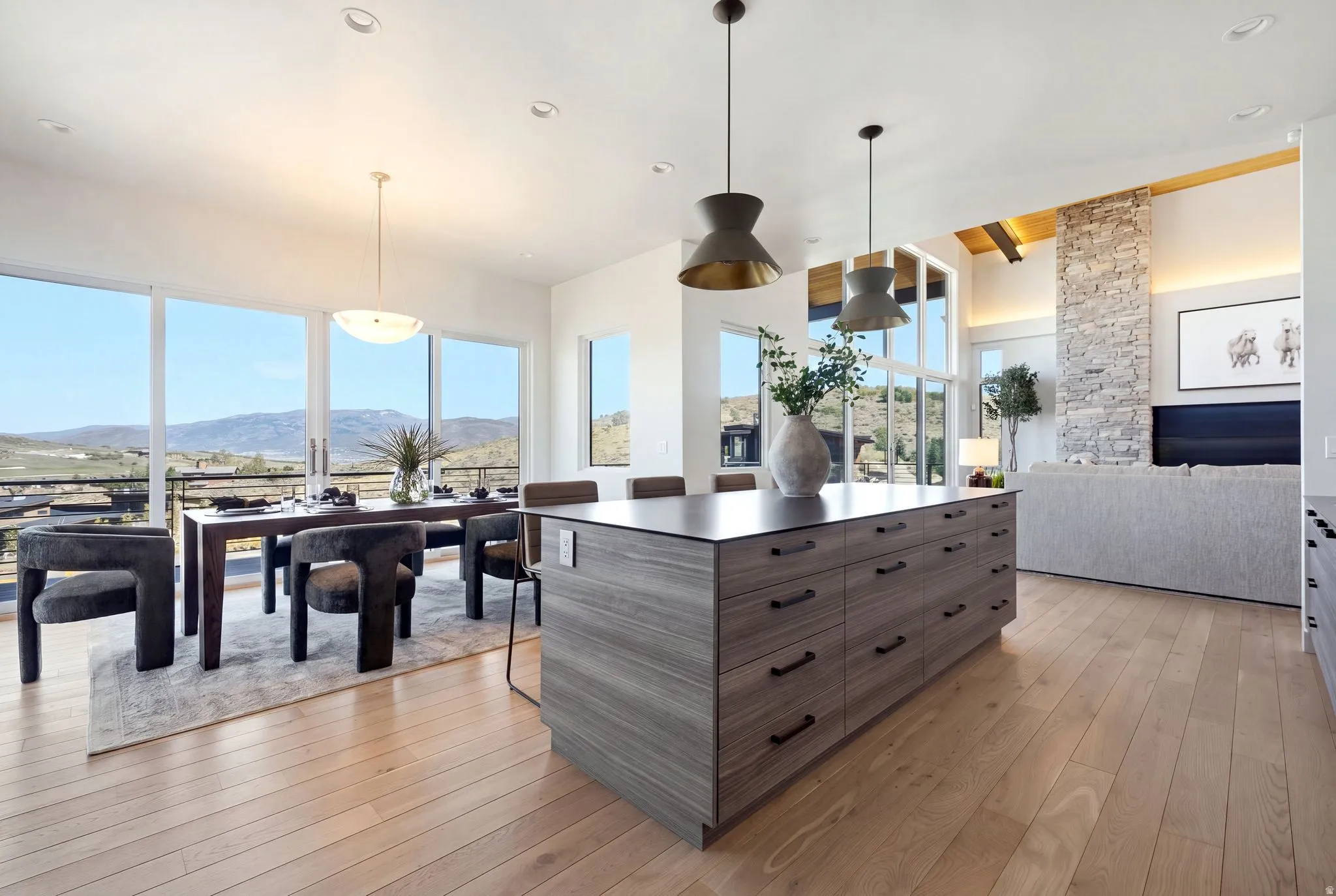 Open-concept living space with light wood-finish flooring, a large kitchen island with dark countertops, and a stone-clad fireplace