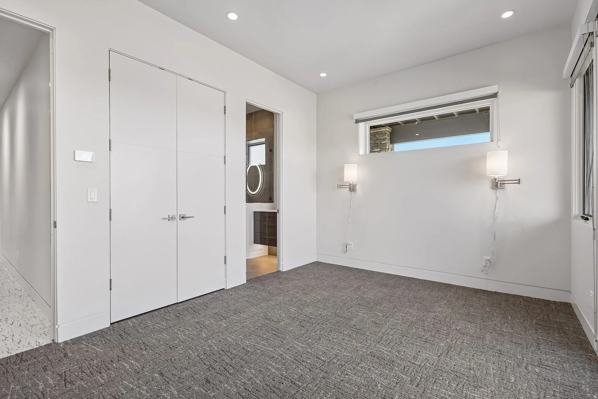 Spacious room featuring patterned gray carpeting, dual white doors, a high-set horizontal window, and two wall-mounted sconces