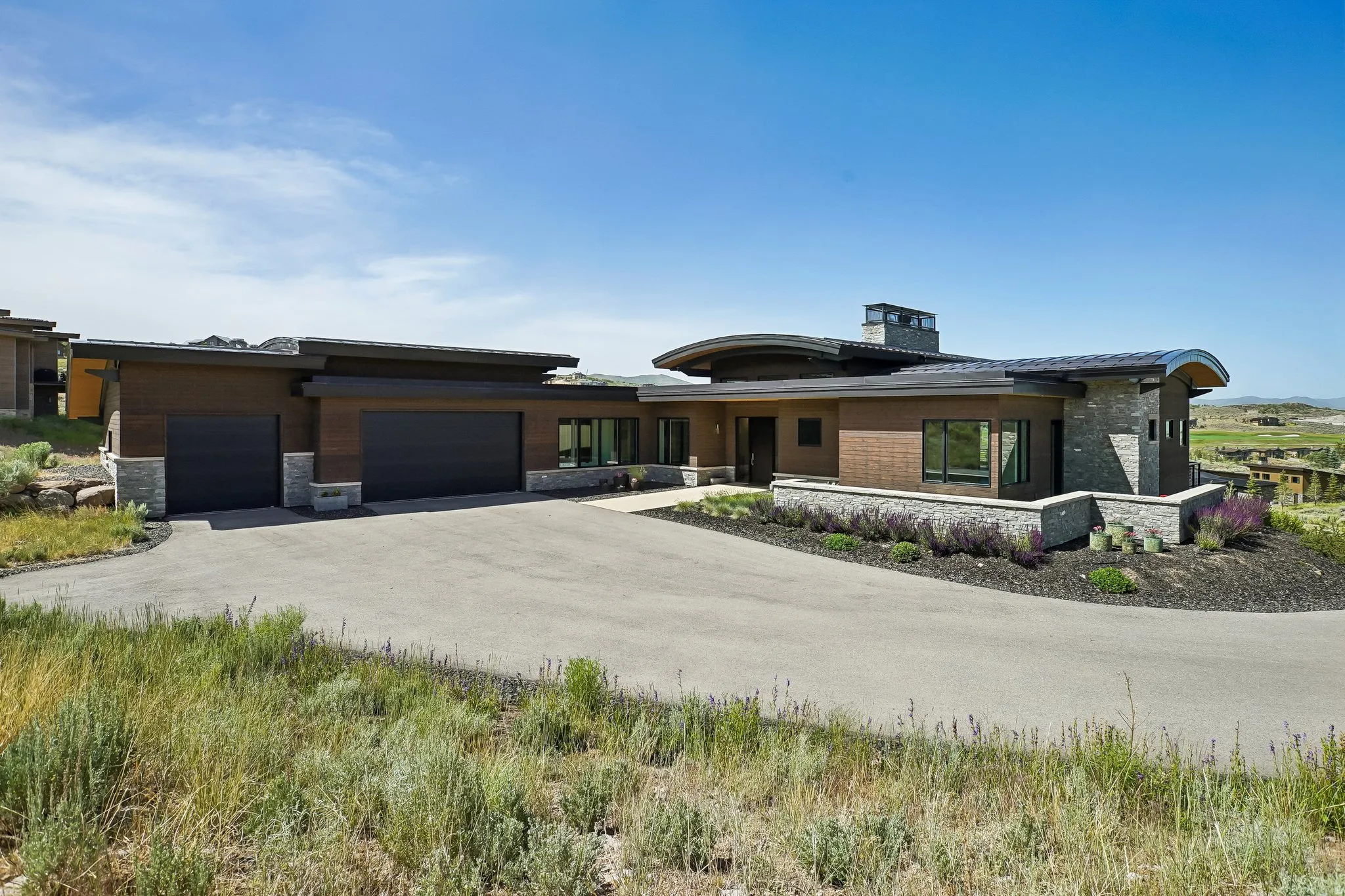 Modern architectural style with a curved roofline, stone and wood-finish exterior, multiple garage doors, extensive paved driveway, and integrated landscaping
