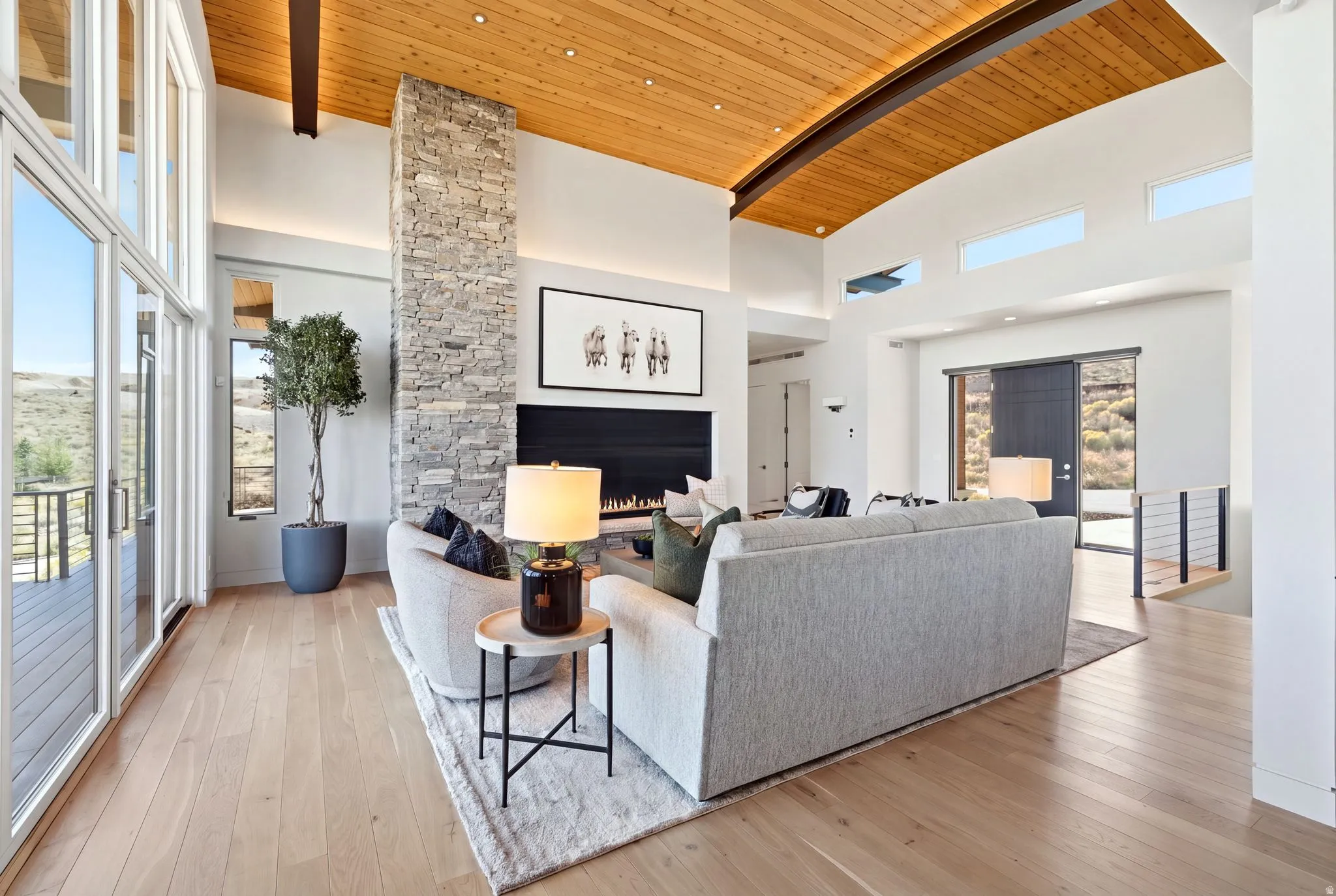 Open-concept living space featuring wood-finish flooring, a stone-clad fireplace, a wood-paneled curved ceiling, and expansive windows