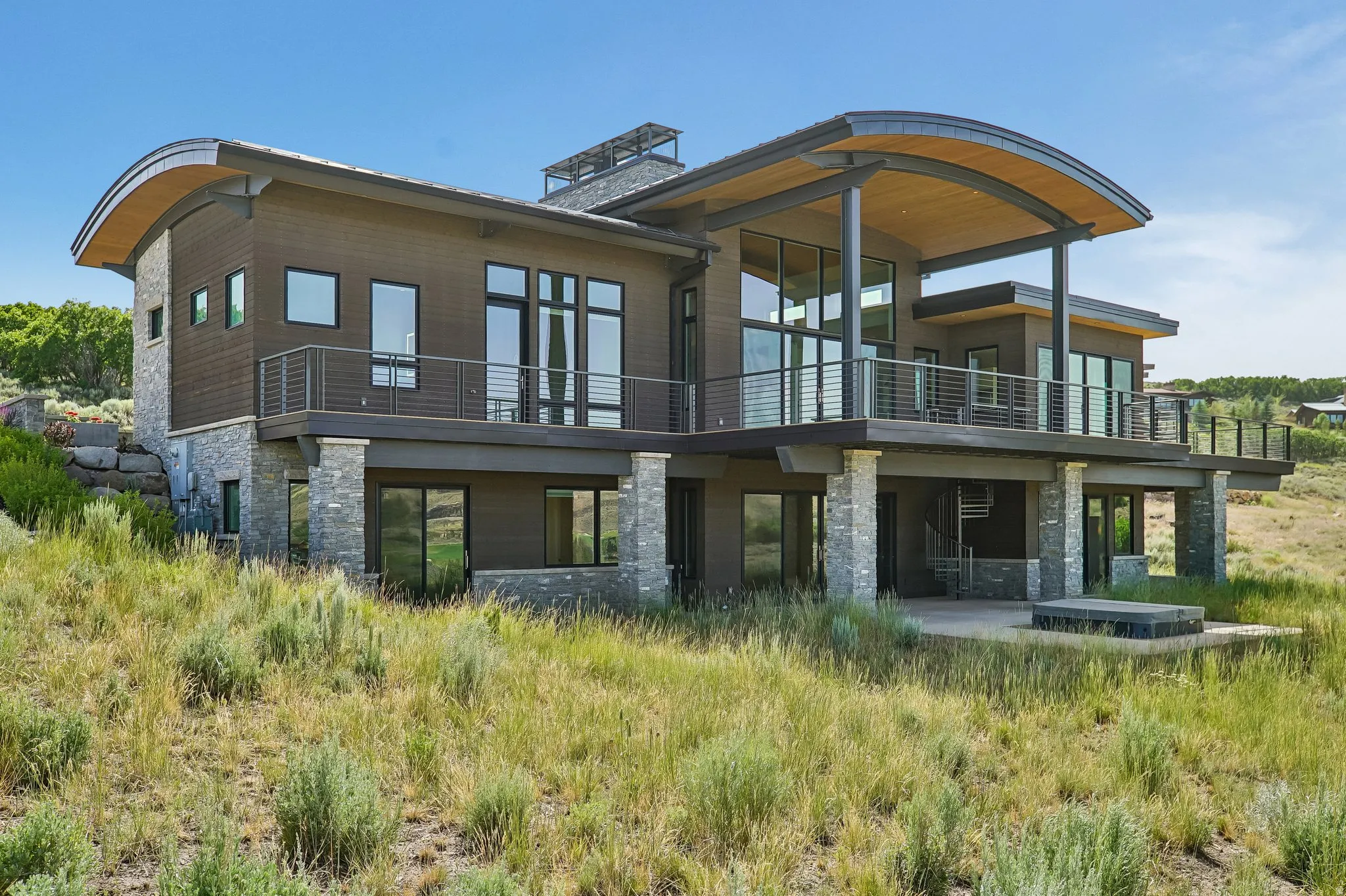 Contemporary architecture featuring a curved wood-paneled soffit, extensive glass windows, multiple balconies with cable railings, stone columns, and a lower-level patio with a built-in fire feature