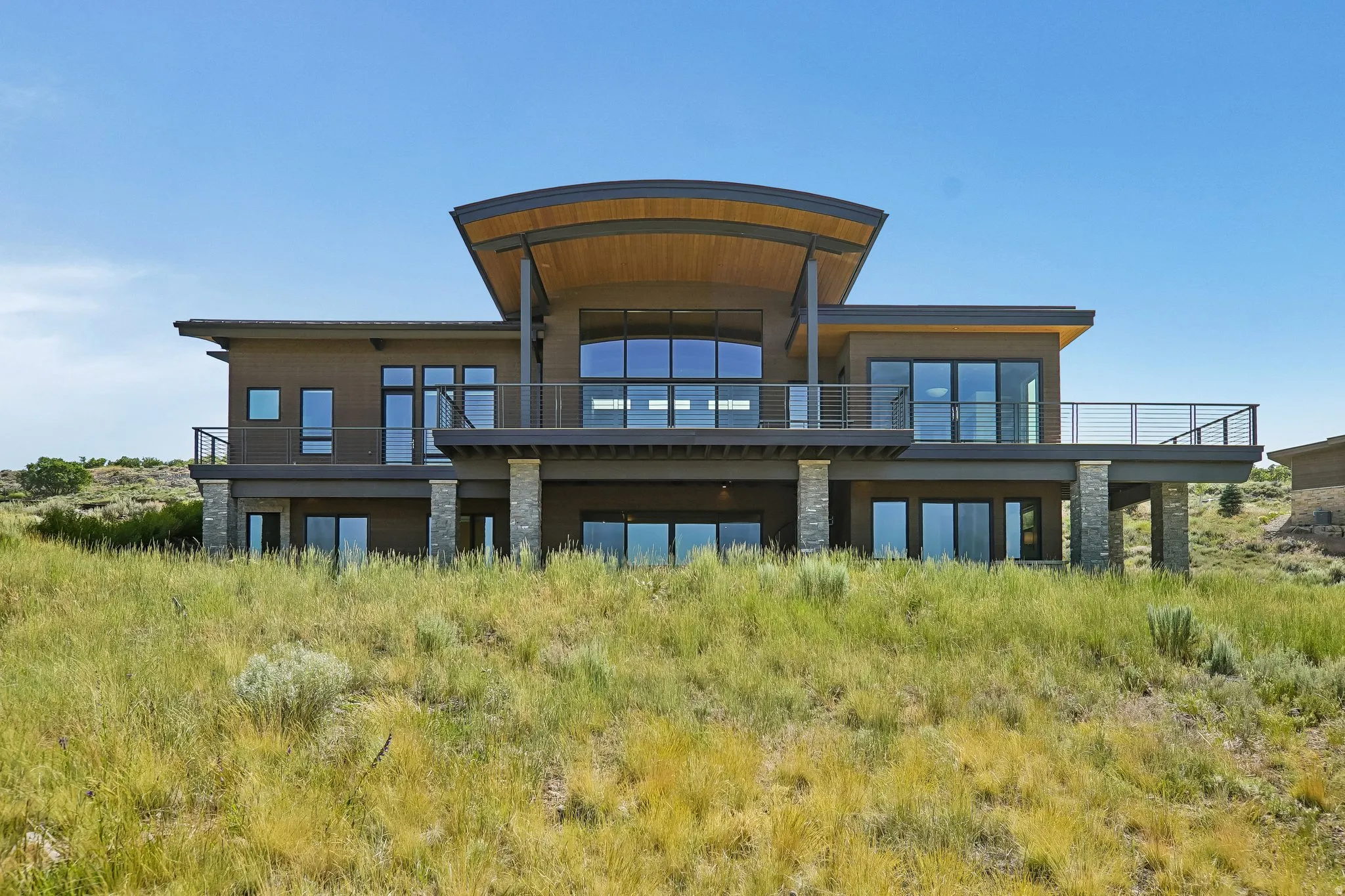 Contemporary architecture featuring a curved roofline, expansive windows, multiple balconies with cable railings, and stone-clad support columns
