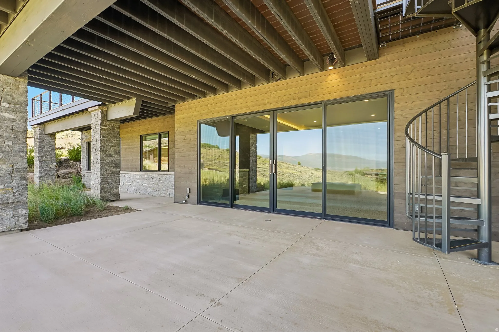 Expansive concrete patio with a spiral metal staircase, large sliding glass doors, stone-clad columns, and a covered ceiling with exposed beams