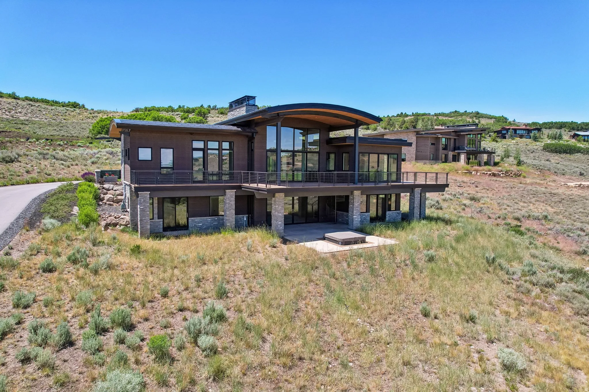 Contemporary residence featuring a curved roofline, expansive glass windows, a large exterior deck with metal railings, a stone-clad foundation, and a ground-level patio with a built-in spa