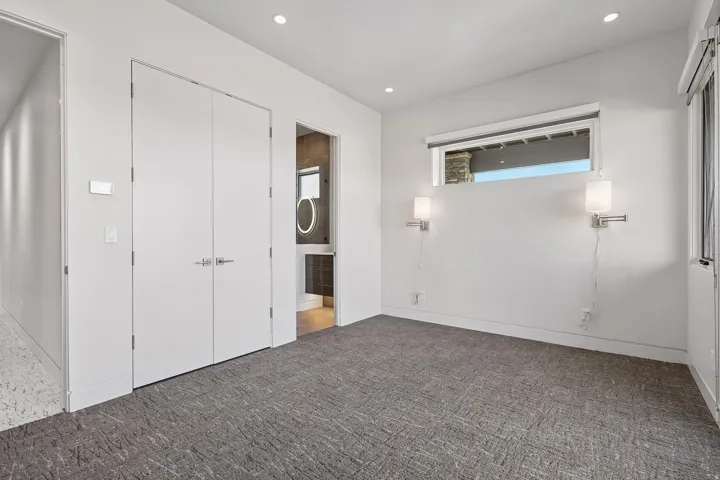 Spacious room featuring patterned gray carpeting, dual white doors, a high-set horizontal window, and two wall-mounted sconces