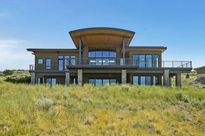Contemporary architecture featuring a curved roofline, expansive windows, multiple balconies with cable railings, and stone-clad support columns