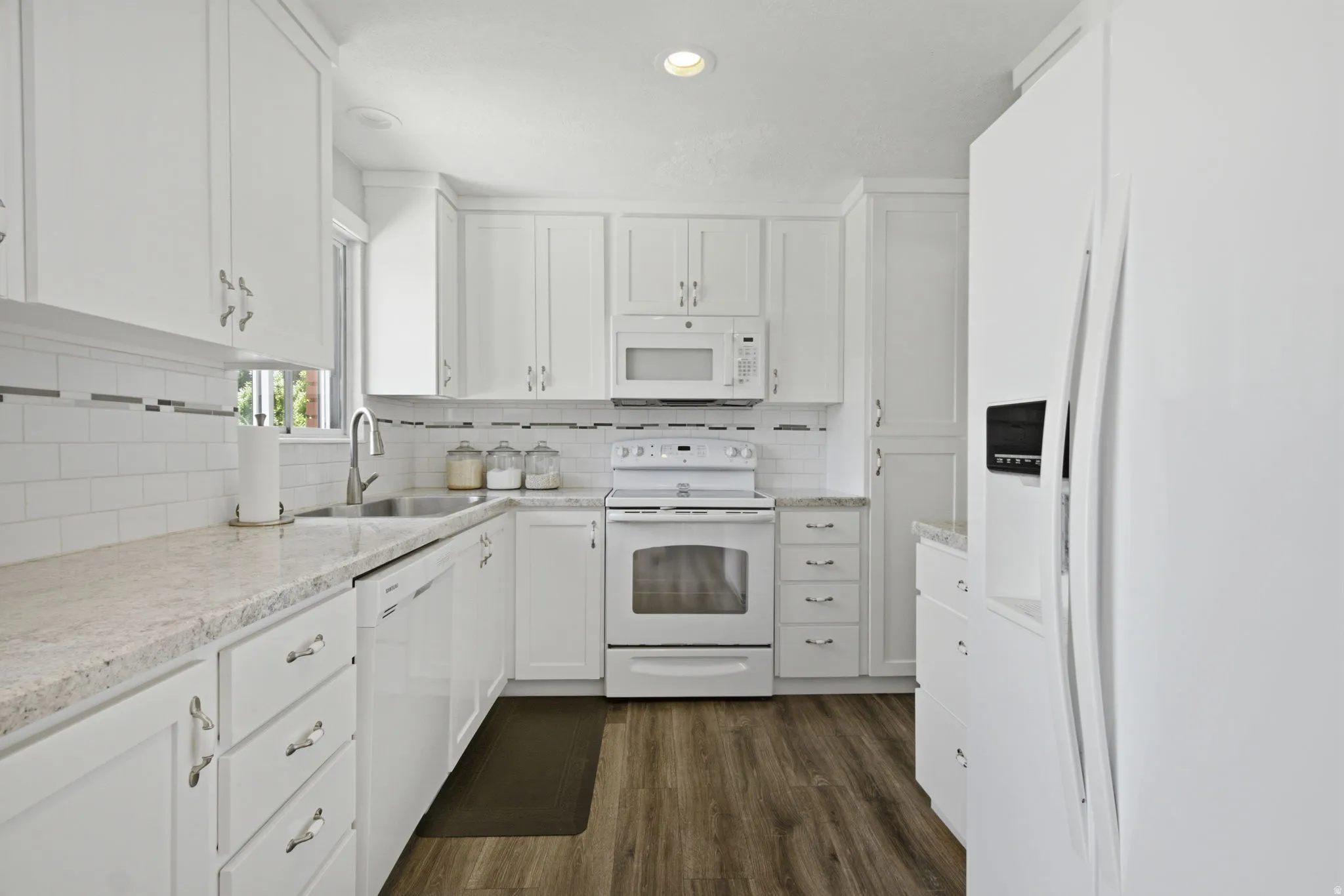 Bright kitchen featuring white cabinetry, light countertops, wood-finish flooring, a stainless steel sink, and white appliances