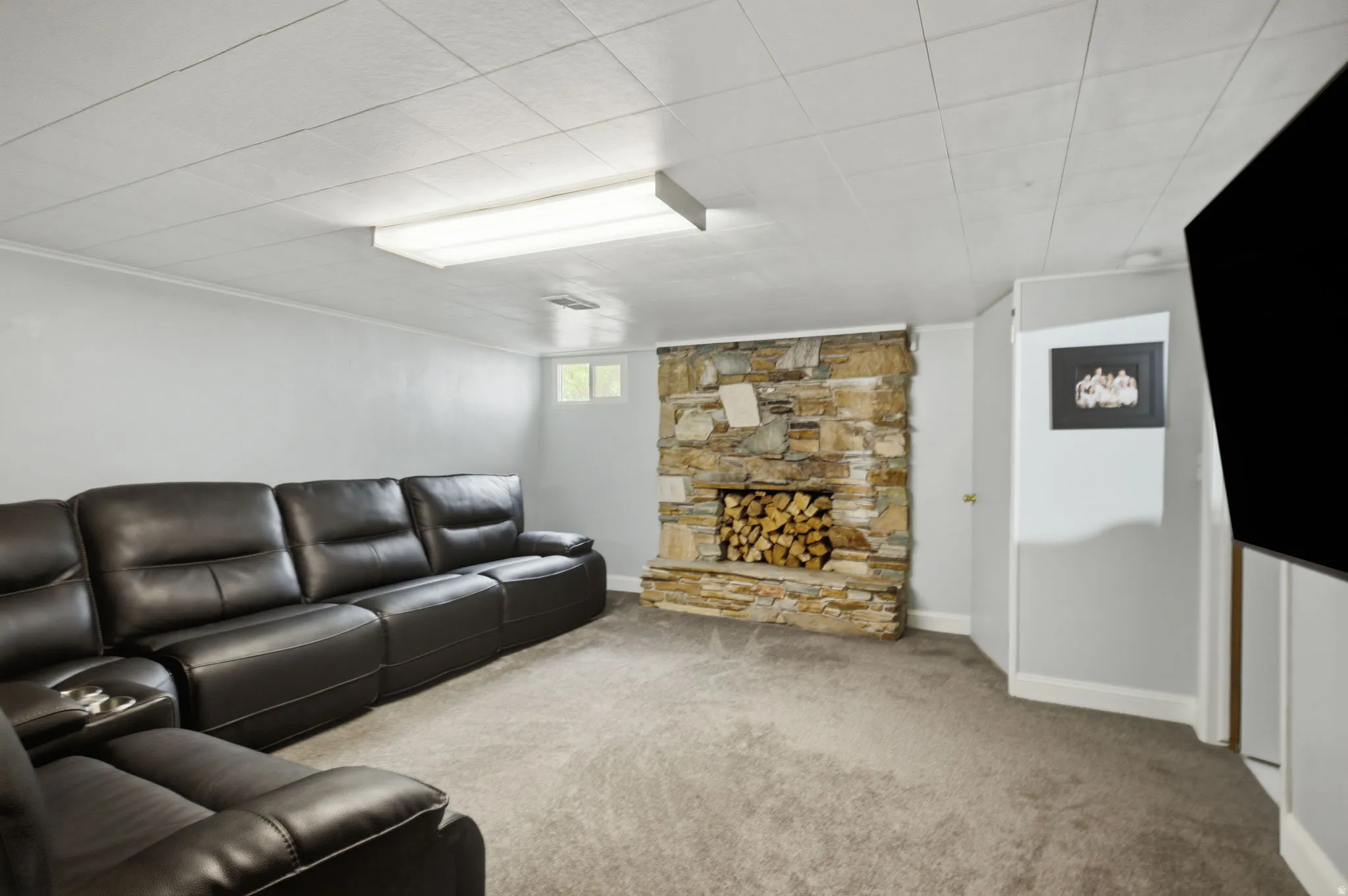 Spacious interior room featuring a stone-clad fireplace with wood storage, light gray walls, gray carpeting, and a drop ceiling with recessed lighting