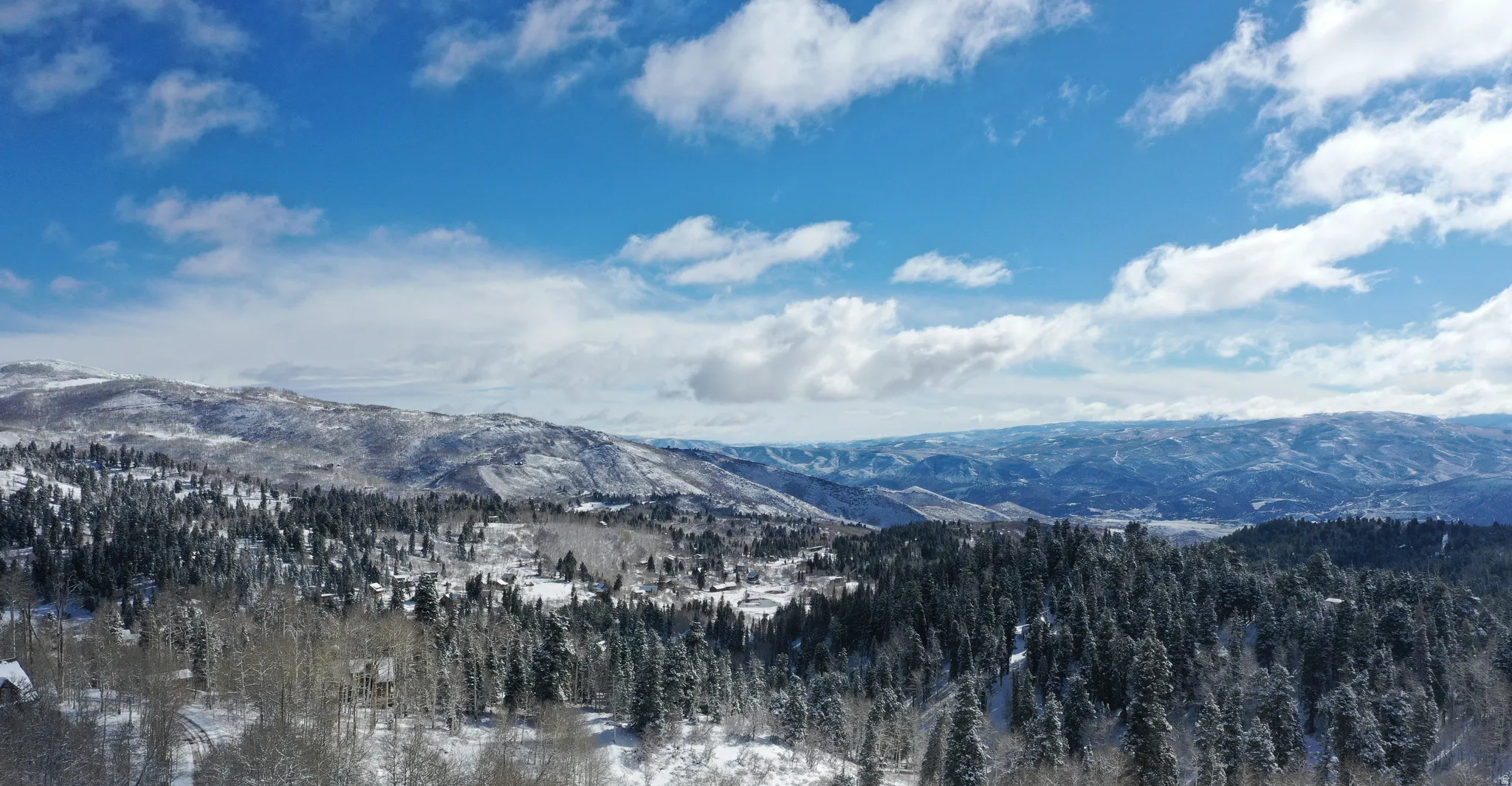 Expansive mountain landscape featuring snow-covered peaks, dense evergreen forests, and a clear blue sky with scattered clouds
