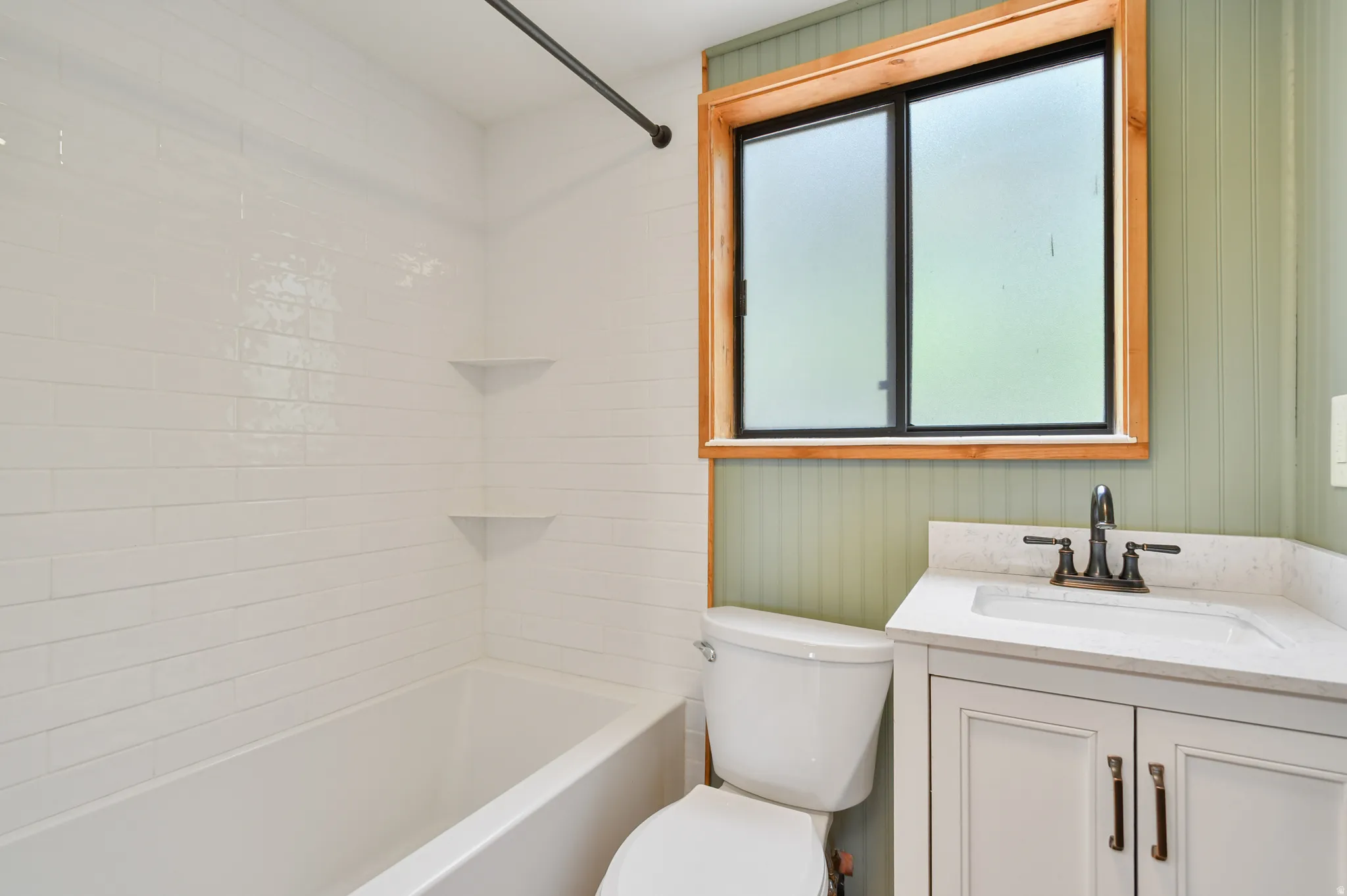 Bathroom featuring a white subway tile tub surround with integrated corner shelves, a natural wood-framed window, and a light green beadboard accent wall