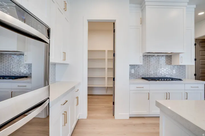 Bright kitchen featuring white cabinetry with gold-finish hardware, stainless steel built-in oven, light wood-finish flooring, and a walk-in pantry with extensive shelving