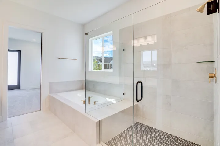 White-tiled bathroom featuring a built-in soaking tub, a frameless glass-enclosed shower with mosaic tile flooring, a window, and brass fixtures