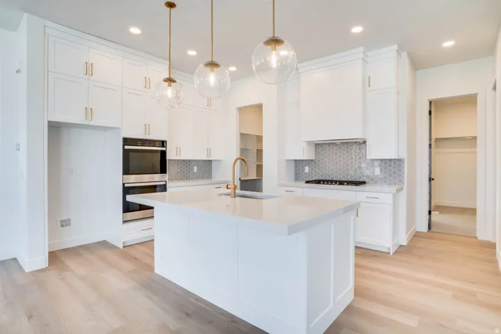 Contemporary kitchen featuring a large central island with an integrated sink, white cabinetry, quartz countertops, a hexagon tile backsplash, and light wood-finish flooring