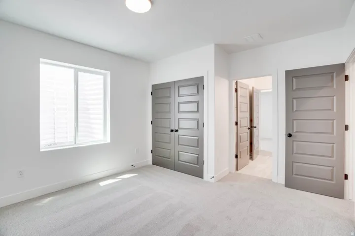 Spacious room featuring light gray carpet, white walls, a large window, a ceiling light fixture, and multiple paneled doors with black hardware