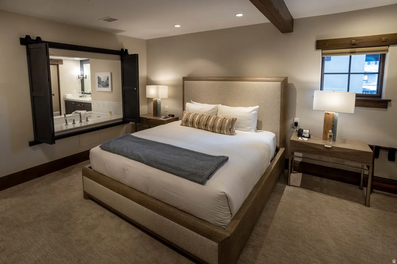 Open concept bedroom featuring a built-in jetted tub with a stone surround, a wood-beam ceiling accent, and custom window shutters