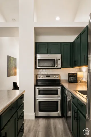 Kitchen featuring dark green cabinetry with matte black hardware, stainless steel appliances, light wood-finish flooring, and light-toned countertops