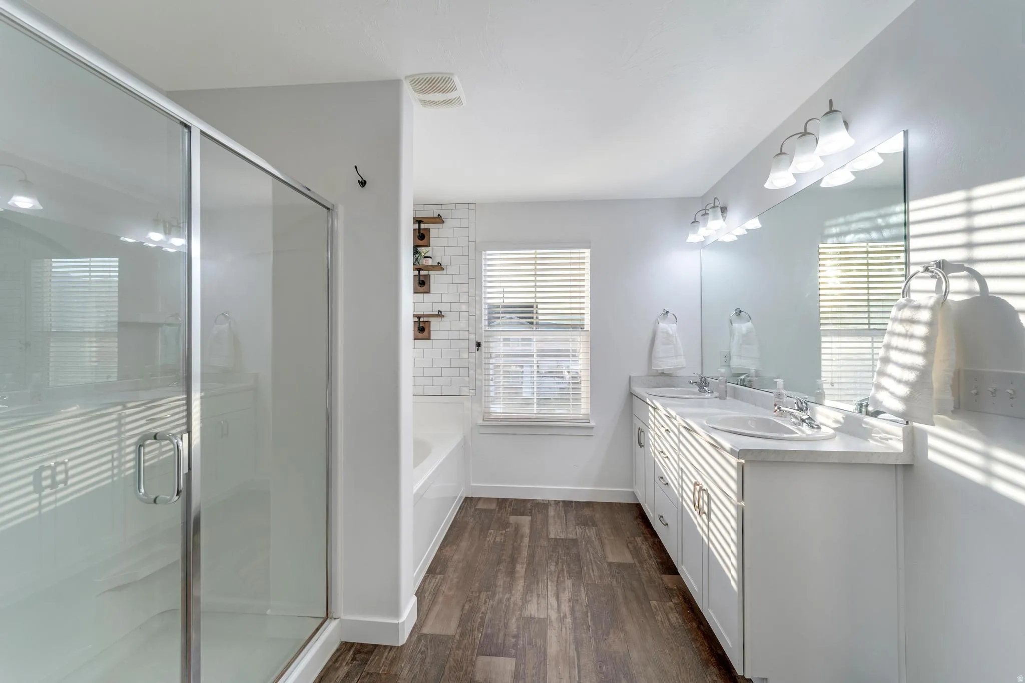 Bathroom featuring a dual vanity with undermount sinks and light-toned countertops, a glass-enclosed shower, a built-in tub with subway tile surround, wood-finish flooring, and a window with horizontal blinds