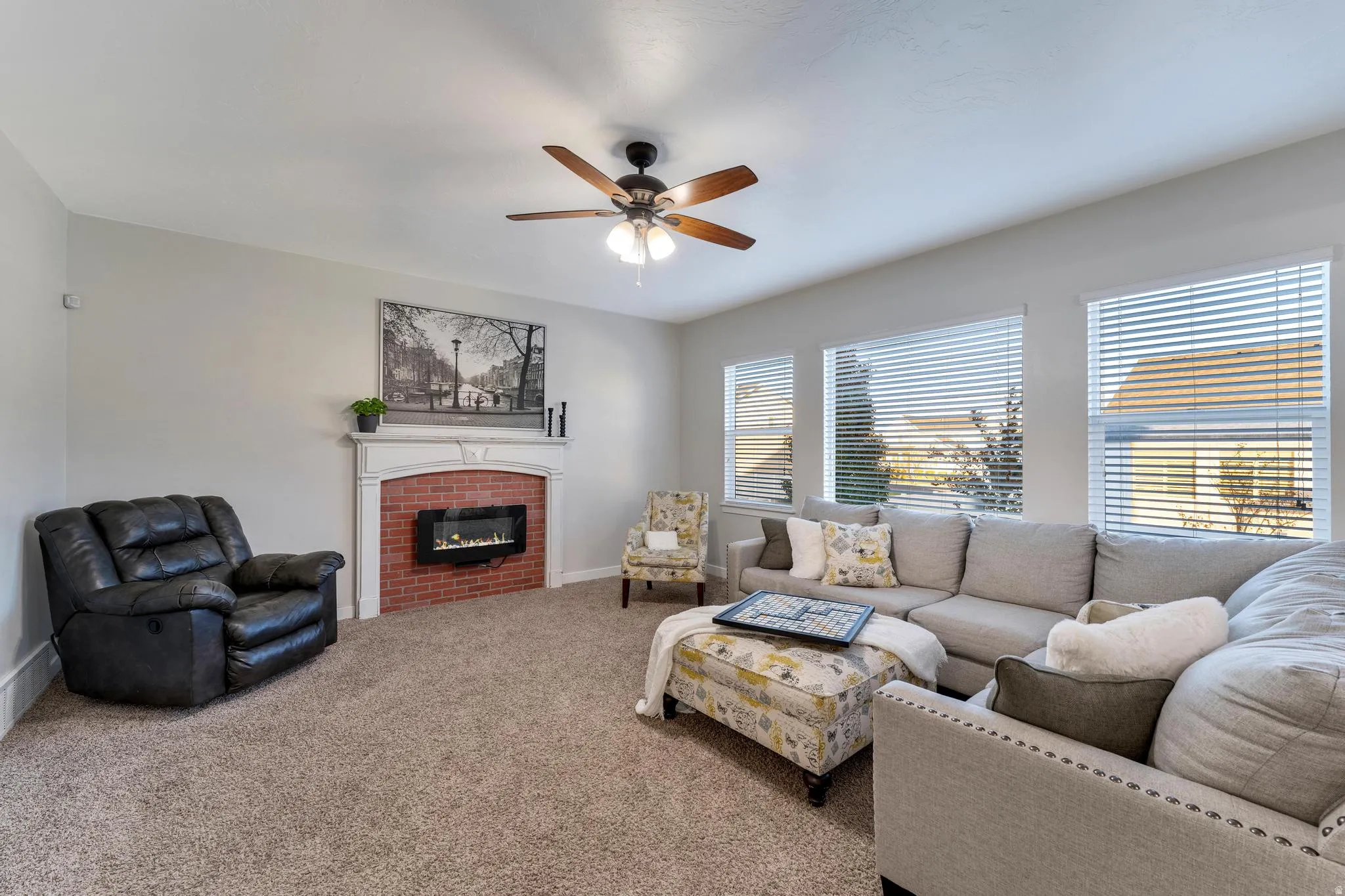 Living area featuring a floor-to-ceiling brick fireplace with a white mantel, ceiling fan with light fixture, and multiple windows