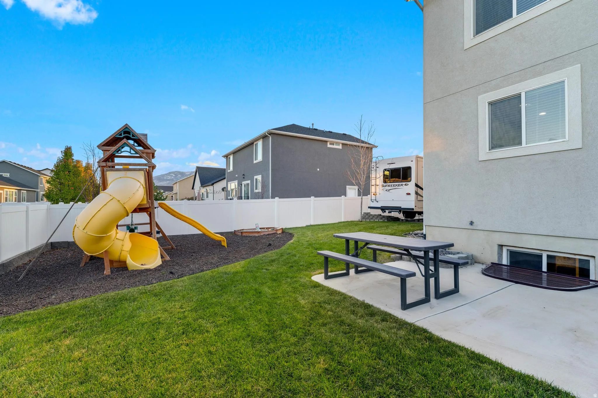 Expansive backyard featuring a manicured lawn, a play structure with slide, a concrete patio area, a picnic table with benches, and a white vinyl privacy fence