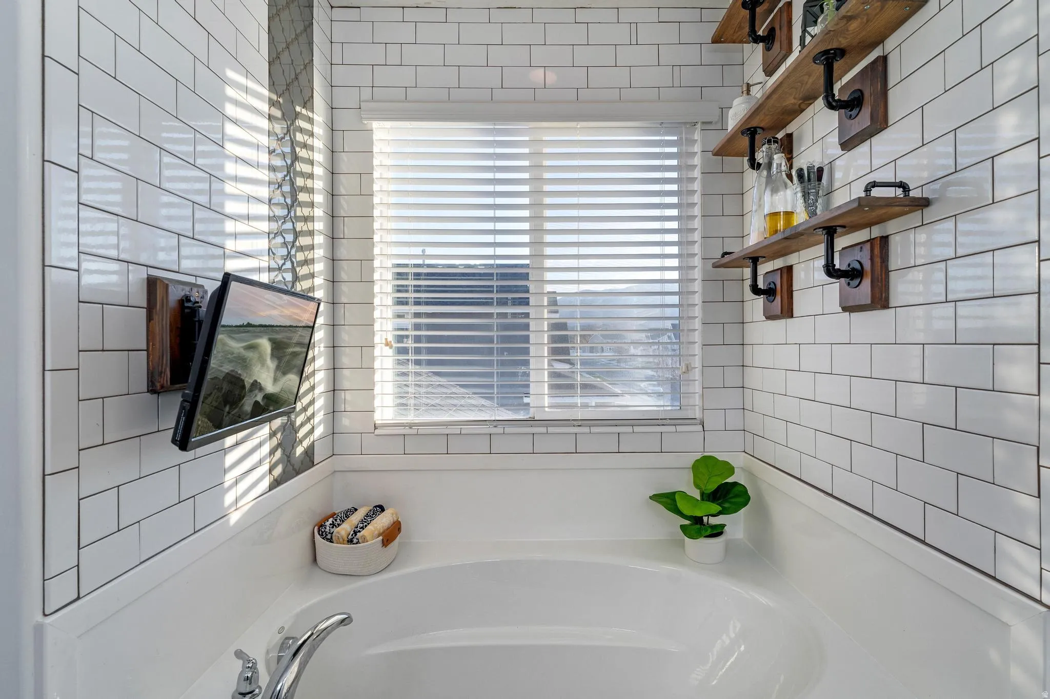 Soaking tub surround with white subway tile, a mounted flat-screen display, and wood-finish floating shelves