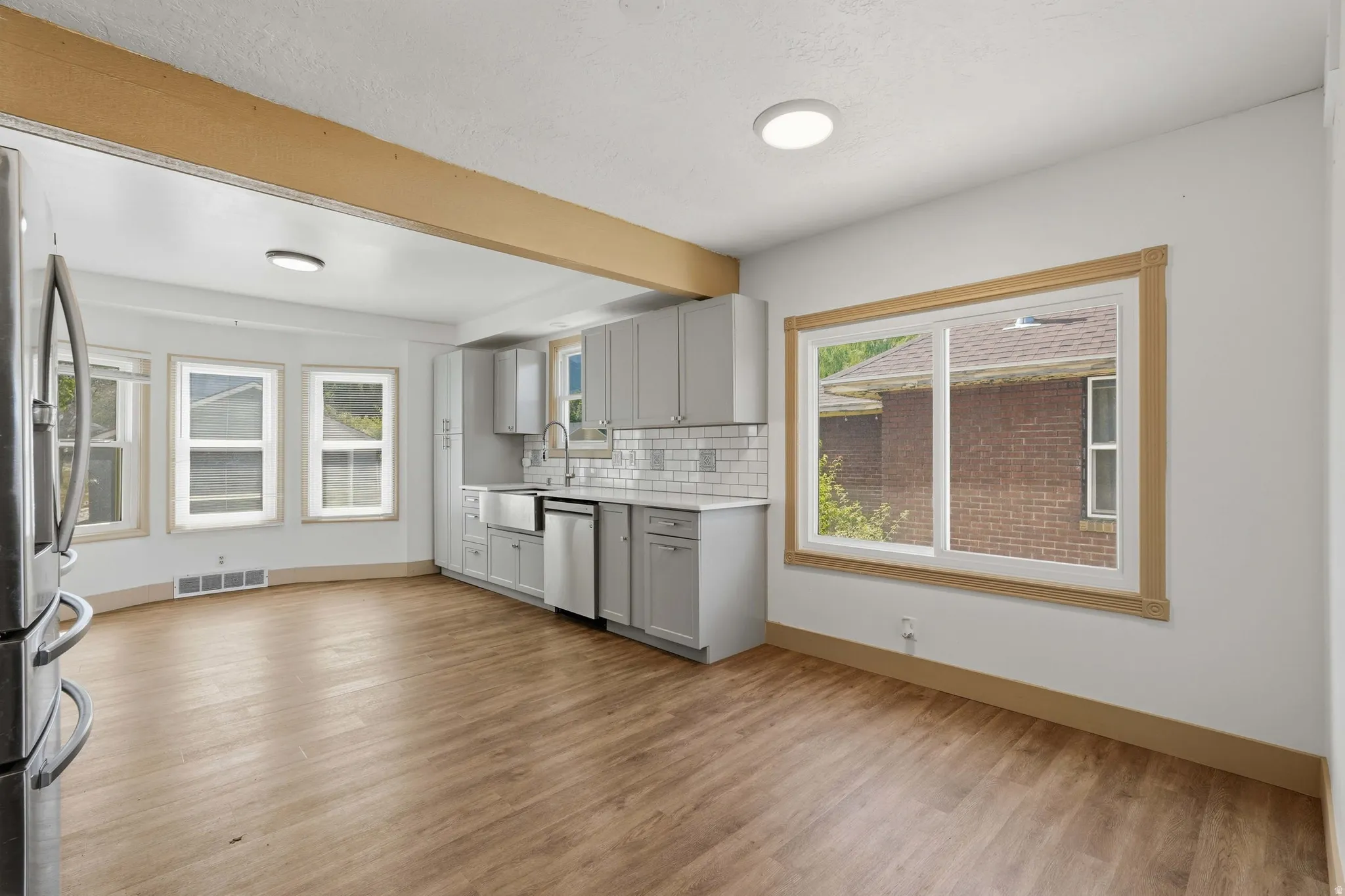 Open-concept kitchen area featuring wood-finish flooring, light gray cabinetry, subway tile backsplash, stainless steel appliances, and multiple windows with wood trim