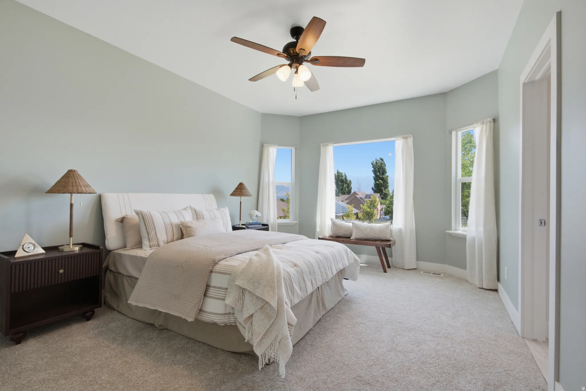 Spacious bedroom featuring light blue walls, soft grey carpeting, a multi-pane window with a deep sill, and a ceiling fan with integrated lighting