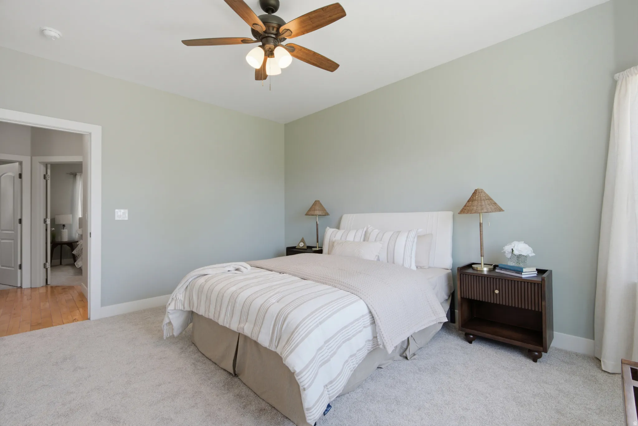 Carpeted bedroom featuring a ceiling fan with light kit, light-colored walls, and an open doorway revealing wood-finish flooring in an adjoining space