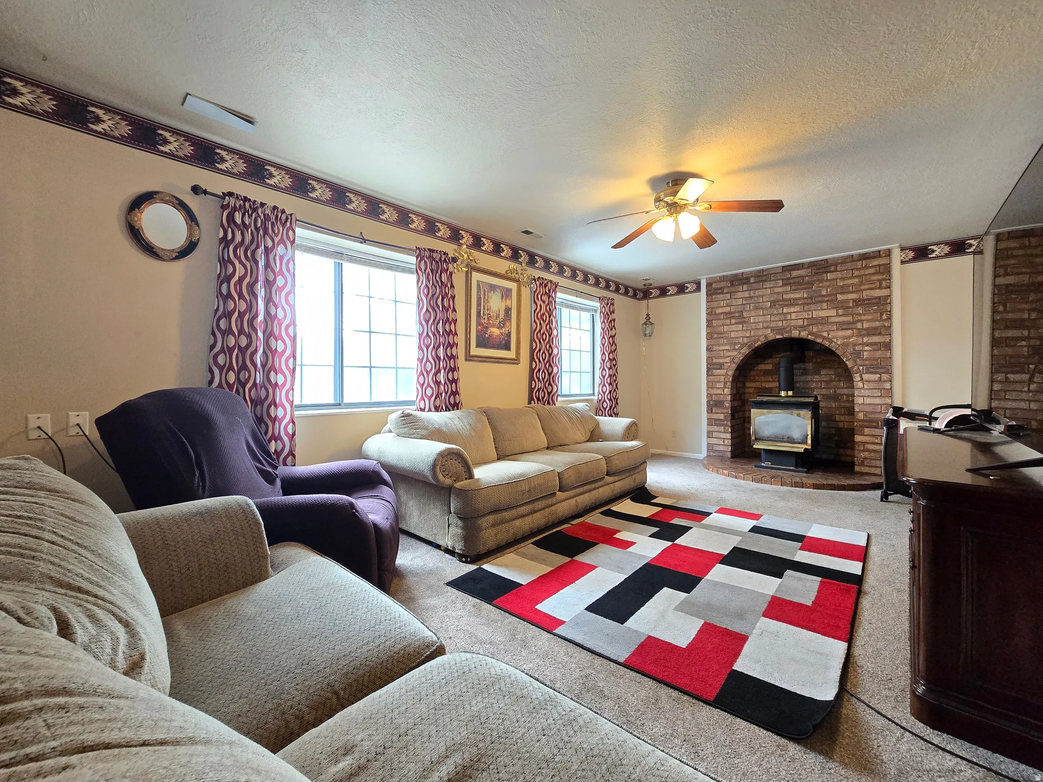 Living area featuring a brick fireplace with an arched opening, a visible wood-burning stove, two windows with grids, ceiling fan with illumination, and wall-to-wall carpeting
