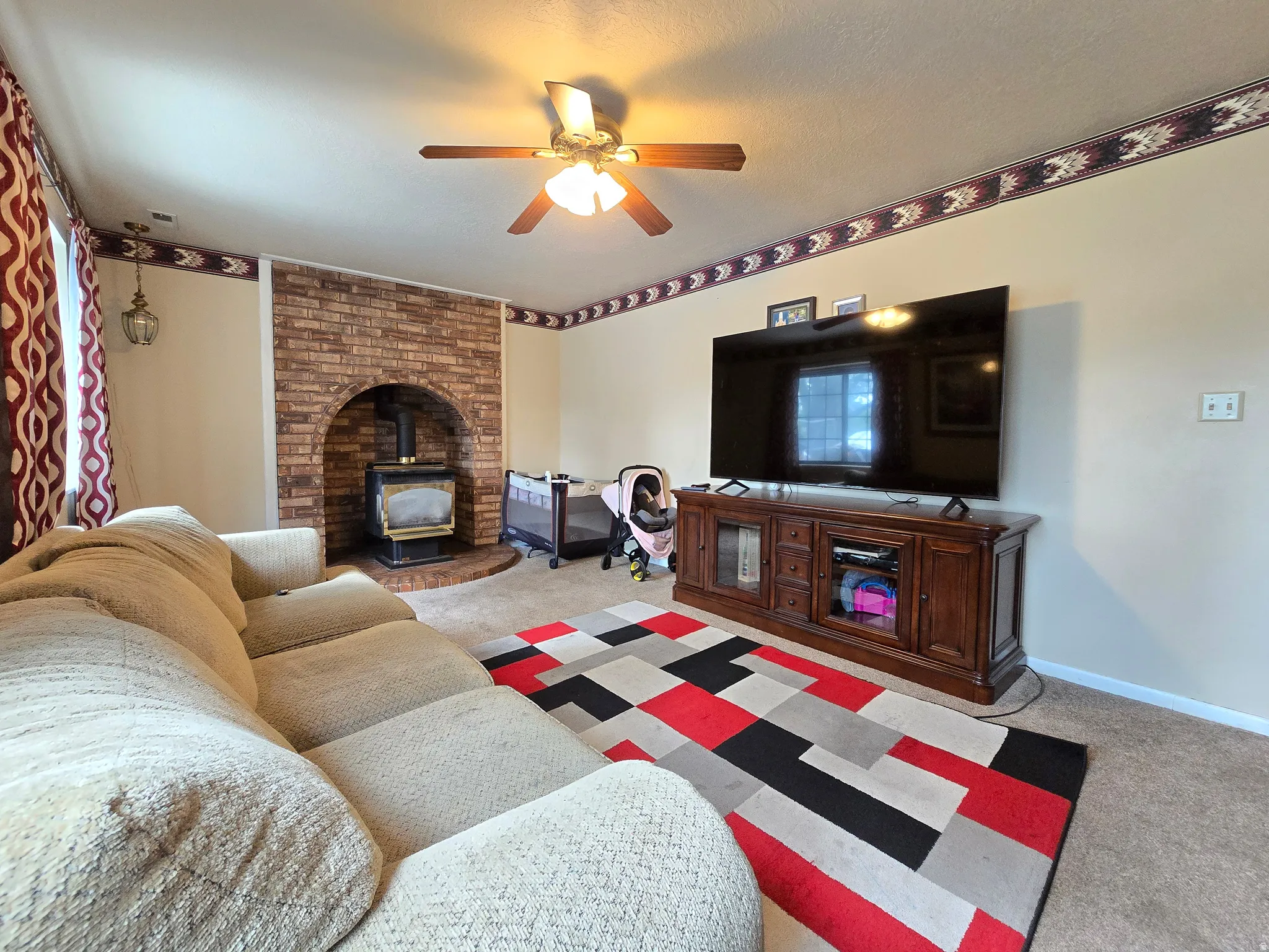 Living room featuring a brick-clad fireplace with wood stove, carpet flooring, a ceiling fan, and wall sconce lighting