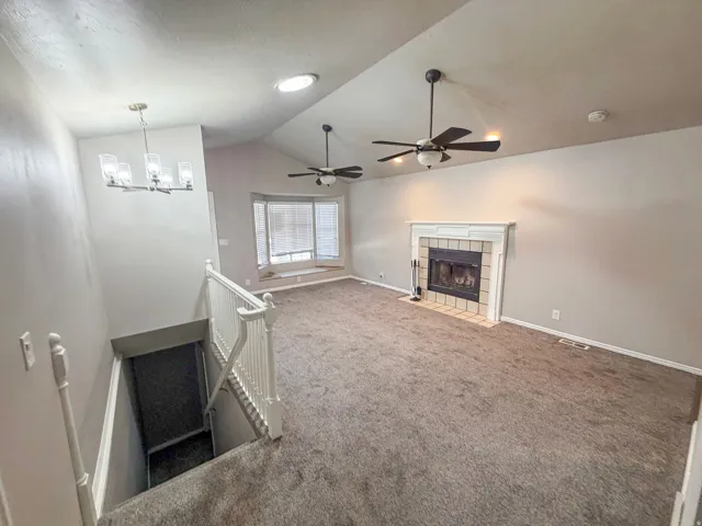 Spacious living area featuring a vaulted ceiling with two ceiling fans, a fireplace with tiled surround and decorative mantel, a bay window with blinds, and plush carpeting