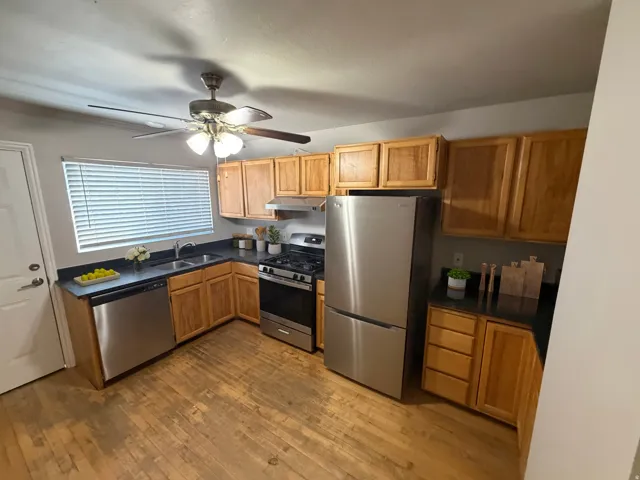 Kitchen featuring wood-finish flooring, natural wood cabinetry, dark countertops, stainless steel appliances, and a ceiling fan with integrated lighting