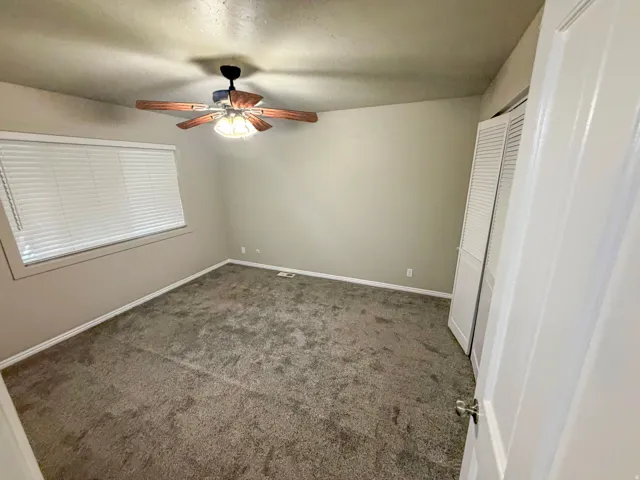 Spacious room featuring neutral carpeting, light beige walls, a window with horizontal blinds, a ceiling fan with a light fixture, and white louvered closet doors