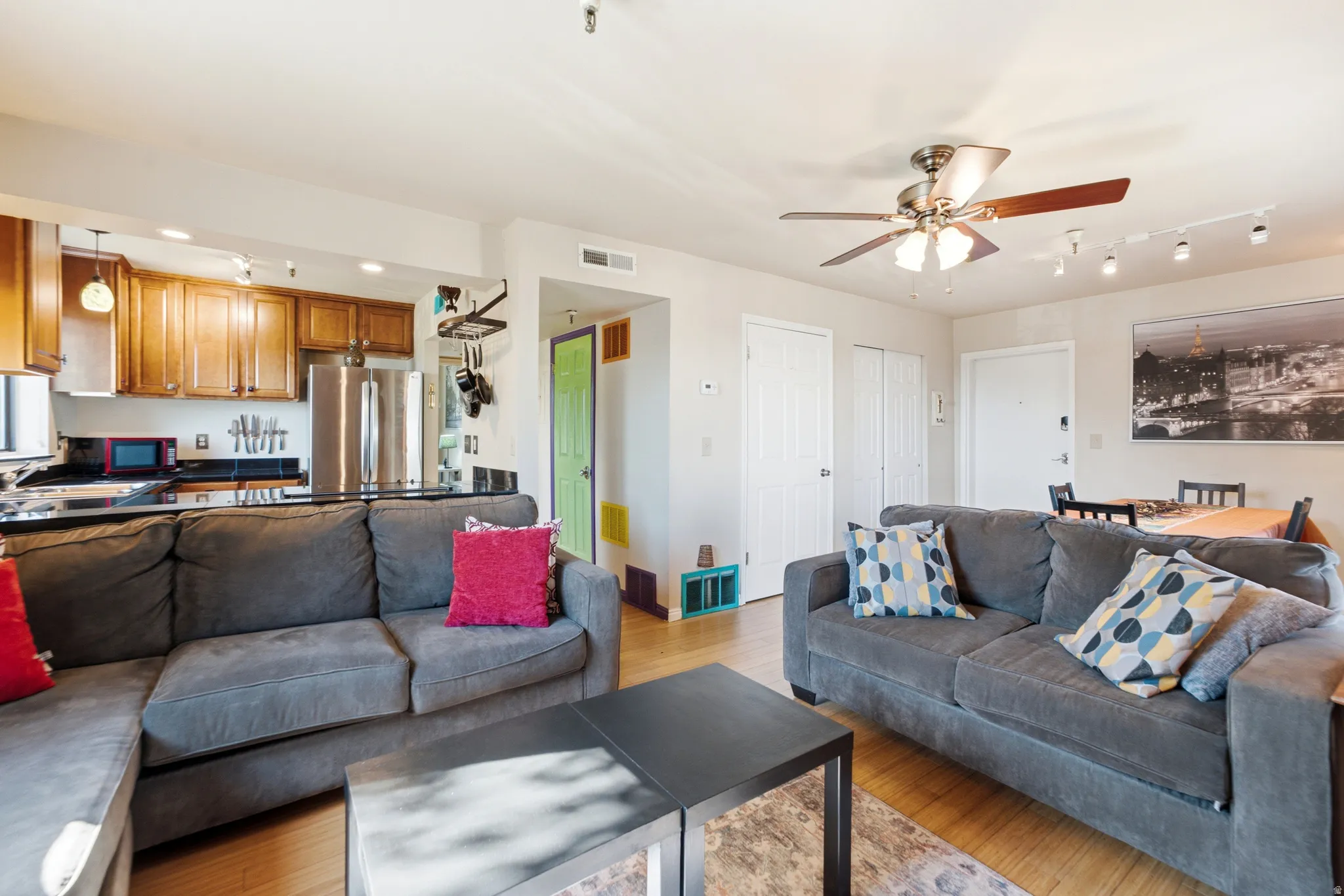 Open-concept living area featuring wood-finish flooring, a kitchen with wood cabinetry and stainless steel appliances, a ceiling fan with integrated lighting, and track lighting