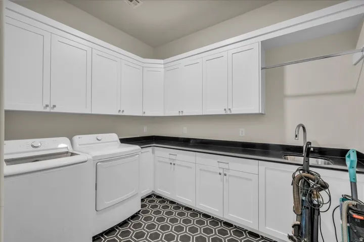 Functional utility room featuring extensive white shaker cabinetry, black countertops, a stainless steel sink with a gooseneck faucet, black and white hexagonal floor tiling, and a built-in hanging rod