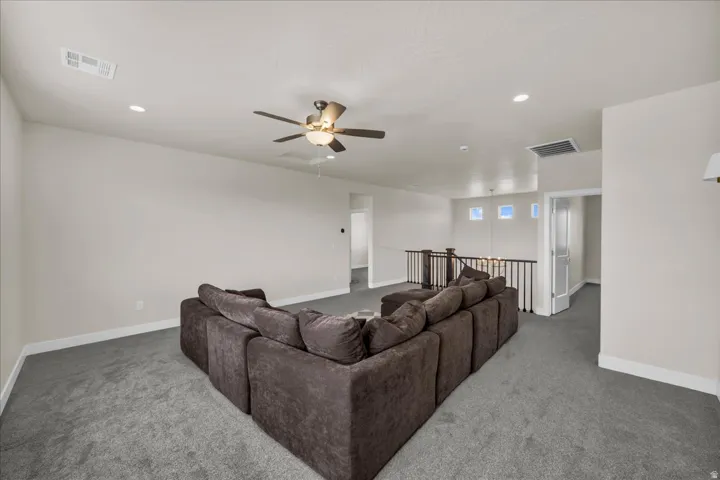Spacious carpeted open area featuring a ceiling fan, recessed lighting, and a dark wood and iron railing