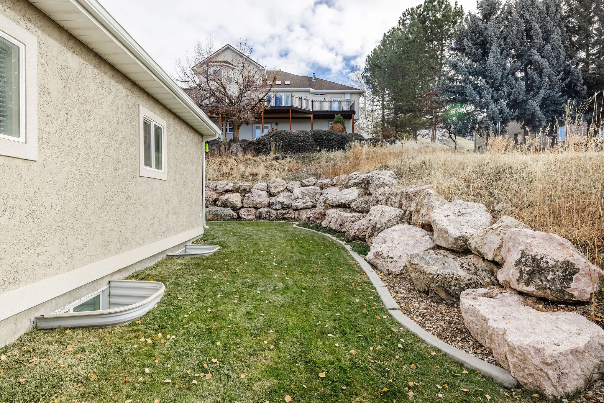 Sloped property featuring a rock retaining wall, mature trees, and a stucco exterior with white-framed windows